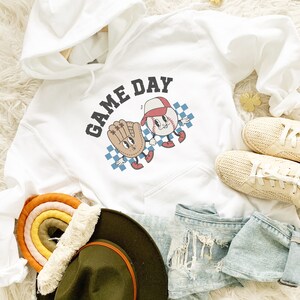 May include: White hoodie with a black graphic of a baseball and a glove with the text "GAME DAY" on it. The hoodie is laying on a white fuzzy surface with a rainbow-colored woven rug, a brown hat, and a pair of white sneakers.