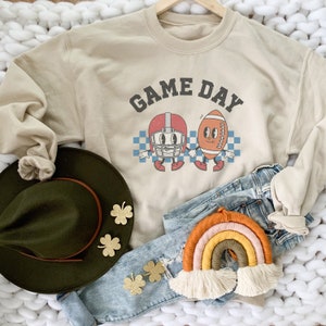 May include: A beige sweatshirt with the text "GAME DAY" in black. The sweatshirt features a cartoon football helmet and football with faces. The sweatshirt is laid out on a white fuzzy surface with a brown hat, blue jeans, and a rainbow-colored macrame hanging decoration.