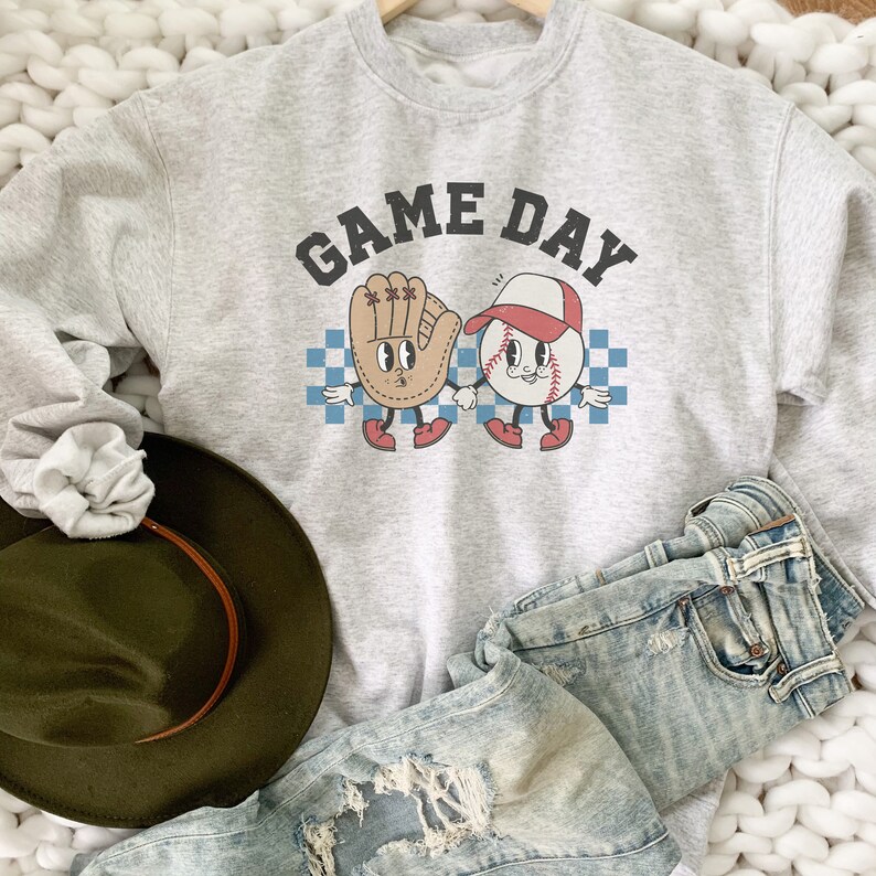 May include: A gray sweatshirt with the text "GAME DAY" in black letters. A cartoon baseball and baseball glove are holding hands. The baseball is wearing a red hat.