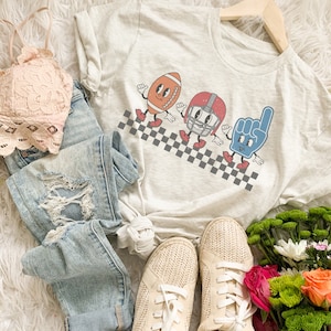 May include: A white t-shirt with a graphic of a football, a helmet, and a foam finger. The graphic is in a retro style with checkered lines behind the characters.