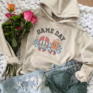 May include: A beige hoodie with a graphic of a football helmet and football with the text "GAME DAY" printed on it. The hoodie is laying on a white fuzzy surface with a pair of blue jeans and a green lace bra.