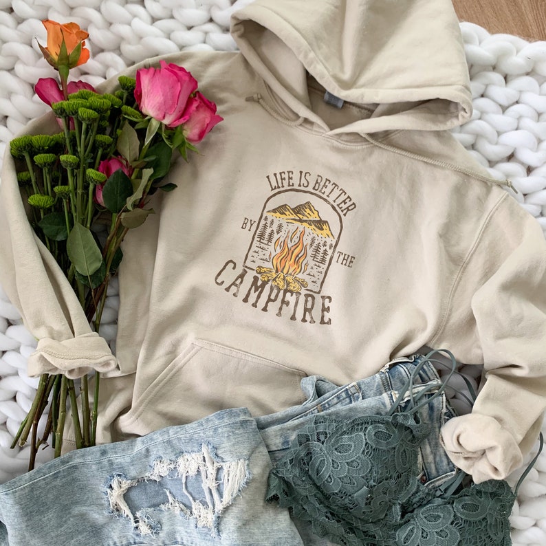 May include: A beige hoodie with the text "LIFE IS BETTER BY THE CAMPFIRE" and a graphic of a campfire, mountains, and trees. The hoodie is paired with light blue jeans, a green lace bralette, and a bouquet of flowers.