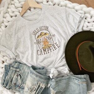 May include: A gray t-shirt with a graphic design that says "Life is better by the campfire". The graphic features a campfire with flames and trees in a brown arch.