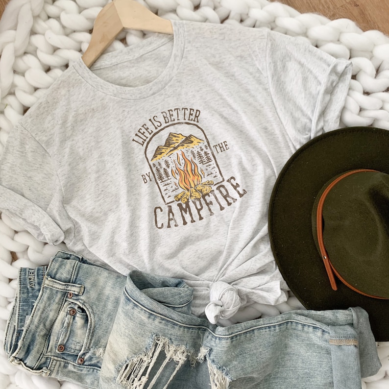 May include: A gray t-shirt with a graphic design that says "Life is better by the campfire". The graphic features a campfire with flames and trees in a brown arch.