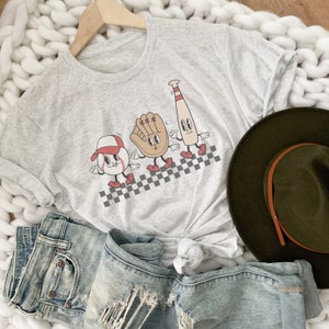 May include: A gray t-shirt with a graphic of a baseball, a baseball glove, and a baseball bat. The graphic is in a cartoon style and the characters are walking on a checkered pattern.