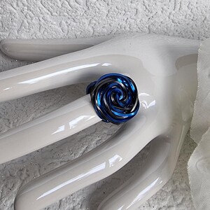 Könnte beinhalten: Ein Ring mit einem spiralförmigen Design in Blau- und Schwarztönen, der einer Rose ähnelt, wird auf einer weißen Hand gezeigt. Das Design des Rings besteht aus geschichteten, metallisch aussehenden Bändern in Blau- und Schwarztönen. Der Hintergrund ist eine strukturierte weiße Oberfläche.