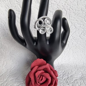 Könnte beinhalten: Ein silberner Ring mit einem wirbelnden, abstrakten Design. Der Ring wird auf einer schwarzen Handpuppe mit einer roten Rose im Vordergrund gezeigt.