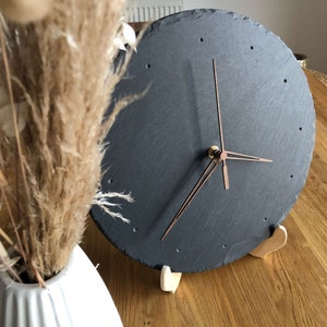 Pode incluir: Um relógio de ardósia preto com ponteiros de cobre e um suporte de madeira. O relógio é redondo e tem um design simples.