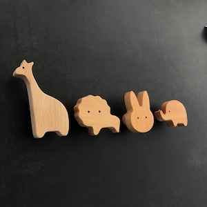 Può includere: Cinque figurine di animali in legno, tra cui una girafa, un leone, un coniglio, un elefante e un albero. Le figurine sono tutte in legno marrone chiaro e hanno caratteristiche semplici e cartoon.