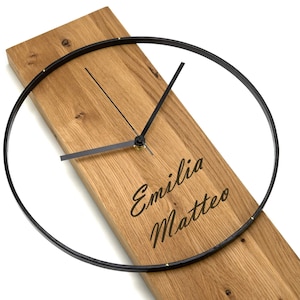 Könnte beinhalten: Eine Holzuhr mit einem schwarzen Metallrahmen und Zeigern. Das Zifferblatt der Uhr ist mit den Namen "Emilia Matteo" graviert.