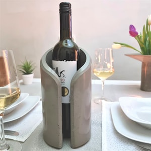 May include: A gray concrete wine bottle holder with a bottle of red wine inside. The holder has a unique design with a curved opening for the bottle.