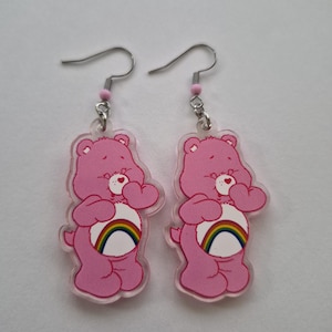 May include: A pair of pink Care Bear earrings. Each earring features a pink bear-shaped charm with a rainbow belly and a heart on its chest. The earrings have silver-colored hooks and small pink beads.