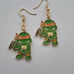 May include: A pair of gold-colored dangle earrings featuring green cartoon turtle characters with orange headbands and holding brown weapons. The earrings have gold-colored fish hook closures.
