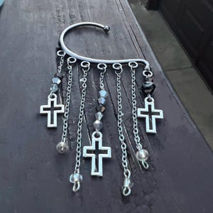 May include: A silver ear cuff with multiple dangling chains and cross charms. Each chain has a small crystal bead and a silver bead.