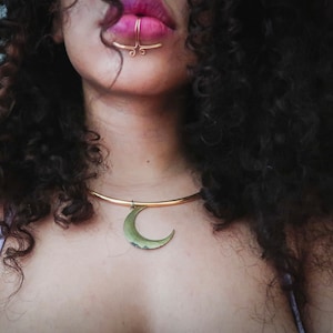 May include: A woman with curly dark hair wearing a gold crescent moon necklace and a gold septum ring.