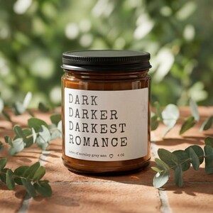 Op de afbeelding: Een bruine glazen potkaars met een zwart deksel. Op het etiket staat "DARK DARKER DARKEST ROMANCE". De kaars is 113 gram en is omgeven door groene bladeren. De pot staat op een bakstenen oppervlak.