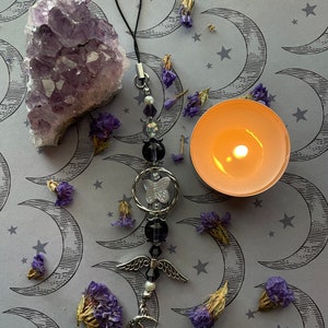 May include: An amethyst crystal cluster, a beaded charm with a butterfly and angel wing design, dried purple flowers, and a lit candle on a gray background with crescent moon and star pattern.