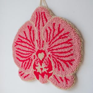 Pode incluir: Uma tape&ccedil;aria de parede tufada rosa e vermelha em forma de flor de orqu&iacute;dea. A flor tem um fundo rosa claro com detalhes vermelhos. As bordas s&atilde;o de cor bege claro. Uma fita bege &eacute; anexada para pendurar.