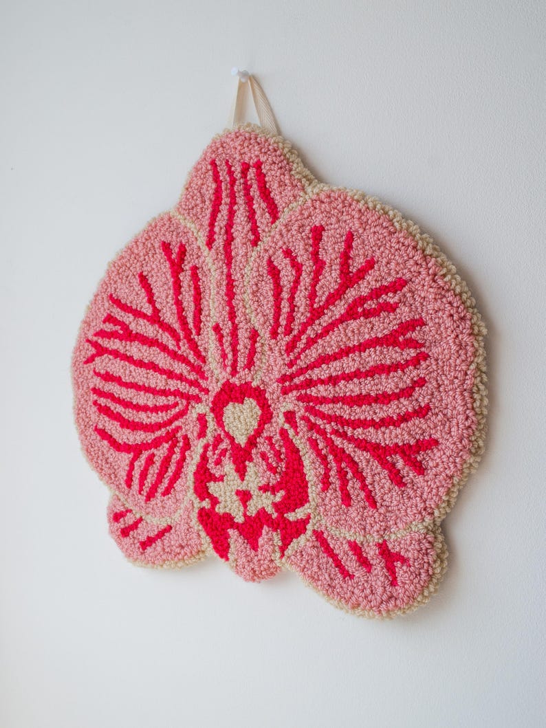 Pode incluir: Uma tape&ccedil;aria de parede tufada rosa e vermelha em forma de flor de orqu&iacute;dea. A flor tem um fundo rosa claro com detalhes vermelhos. As bordas s&atilde;o de cor bege claro. Uma fita bege &eacute; anexada para pendurar.