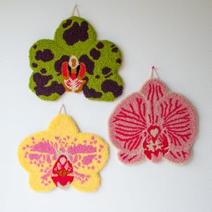 Pode incluir: Tr&ecirc;s penduradores de parede coloridos em forma de flores de orqu&iacute;dea. Os desenhos tufados apresentam detalhes em vermelho, roxo e branco. Cada flor &eacute; suspensa em uma parede branca com um la&ccedil;o para pendurar.