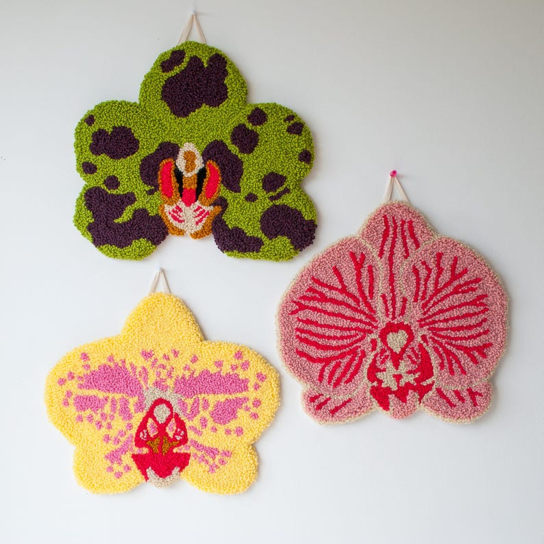 Pode incluir: Tr&ecirc;s penduradores de parede coloridos em forma de flores de orqu&iacute;dea. Os desenhos tufados apresentam detalhes em vermelho, roxo e branco. Cada flor &eacute; suspensa em uma parede branca com um la&ccedil;o para pendurar.