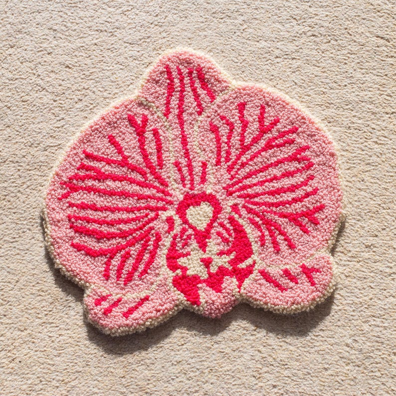 Pode incluir: Um tapete tufado rosa e vermelho em forma de flor de orqu&iacute;dea. O tapete apresenta um fundo rosa claro com um design floral vermelho detalhado. O tapete &eacute; colocado sobre um tapete bege.