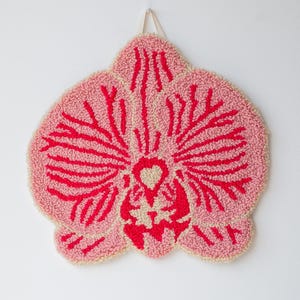 Pode incluir: Uma tape&ccedil;aria de parede tufada rosa e vermelha em forma de flor de orqu&iacute;dea. A flor tem um fundo rosa claro com detalhes vermelhos e um centro creme. Uma fita branca &eacute; anexada para pendurar.