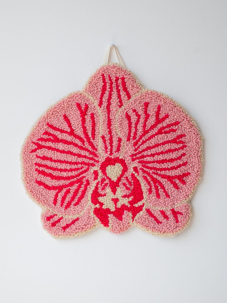 Pode incluir: Uma tape&ccedil;aria de parede tufada rosa e vermelha em forma de flor de orqu&iacute;dea. A flor tem um fundo rosa claro com detalhes vermelhos e um centro creme. Uma fita branca &eacute; anexada para pendurar.