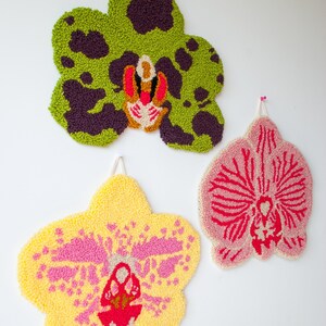 Pode incluir: Tr&ecirc;s tape&ccedil;arias de parede de orqu&iacute;deas tufadas e coloridas. A superior &eacute; verde com manchas roxas escuras. A do meio &eacute; amarela com detalhes em rosa, e a inferior &eacute; rosa com detalhes em vermelho. Cada uma tem uma argola para pendurar.