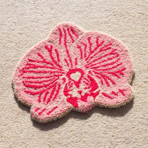 Pode incluir: Um tapete rosa e vermelho em forma de flor com um design texturizado e tufado. O tapete apresenta uma base rosa claro com um padr&atilde;o floral vermelho vibrante. O tapete &eacute; colocado sobre uma superf&iacute;cie de carpete bege.