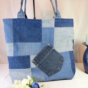 May include: A patchwork denim tote bag with a pocket detail. The bag is constructed from various shades of blue denim, creating a textured, geometric pattern. It has denim handles and a rectangular shape, suitable for carrying items.