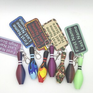 May include: A collection of seven bowling pin keychains with metal lobster clasps, each paired with a rectangular tag featuring the text First Name Last Name and various designs including flames, camouflage, and solid colors.