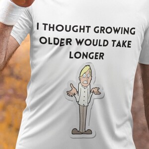 Puede incluir: Camiseta blanca con un gráfico negro que dice "Pensé que envejecer llevaría más tiempo". El gráfico también incluye una ilustración de dibujos animados de una persona con una expresión confundida.