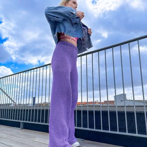 May include: A woman wearing a light blue denim jacket, a pink bra, and wide-leg, lavender knit trousers. She is standing on a wooden deck with a metal railing in the background.