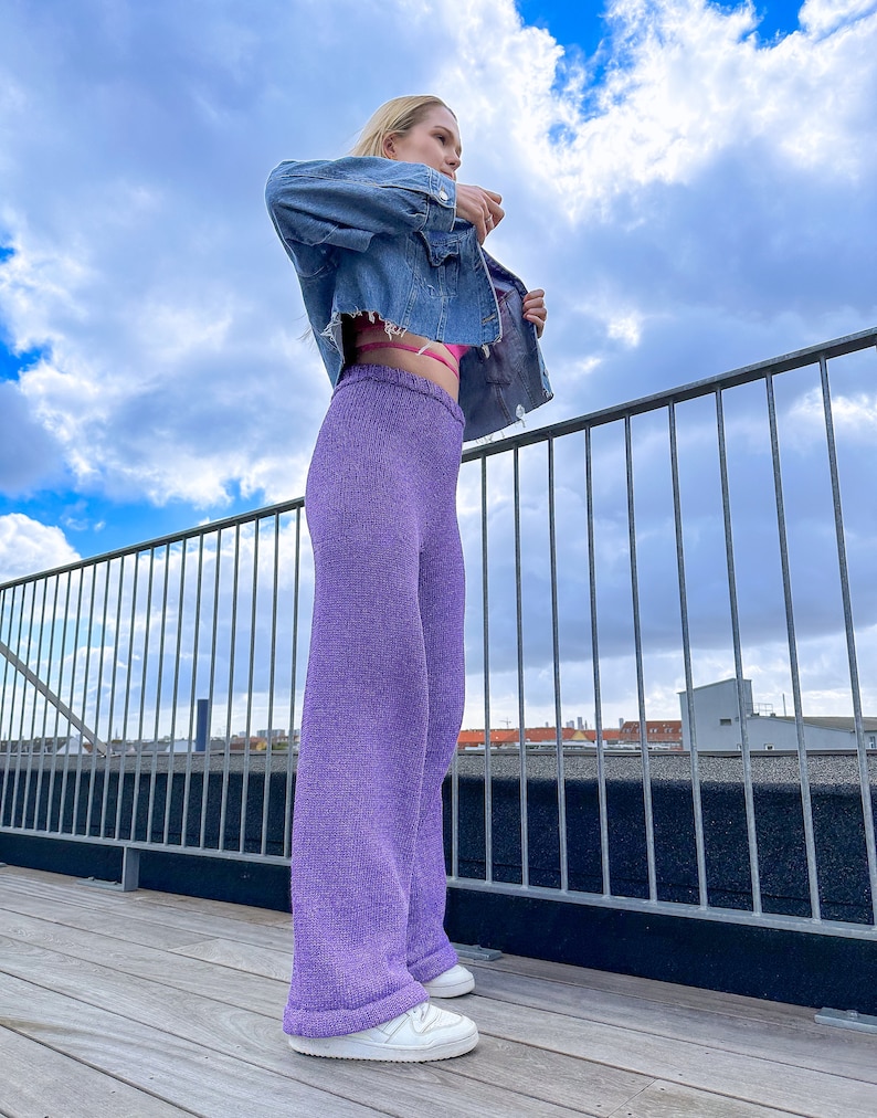 May include: A woman wearing a light blue denim jacket, a pink bra, and wide-leg, lavender knit trousers. She is standing on a wooden deck with a metal railing in the background.