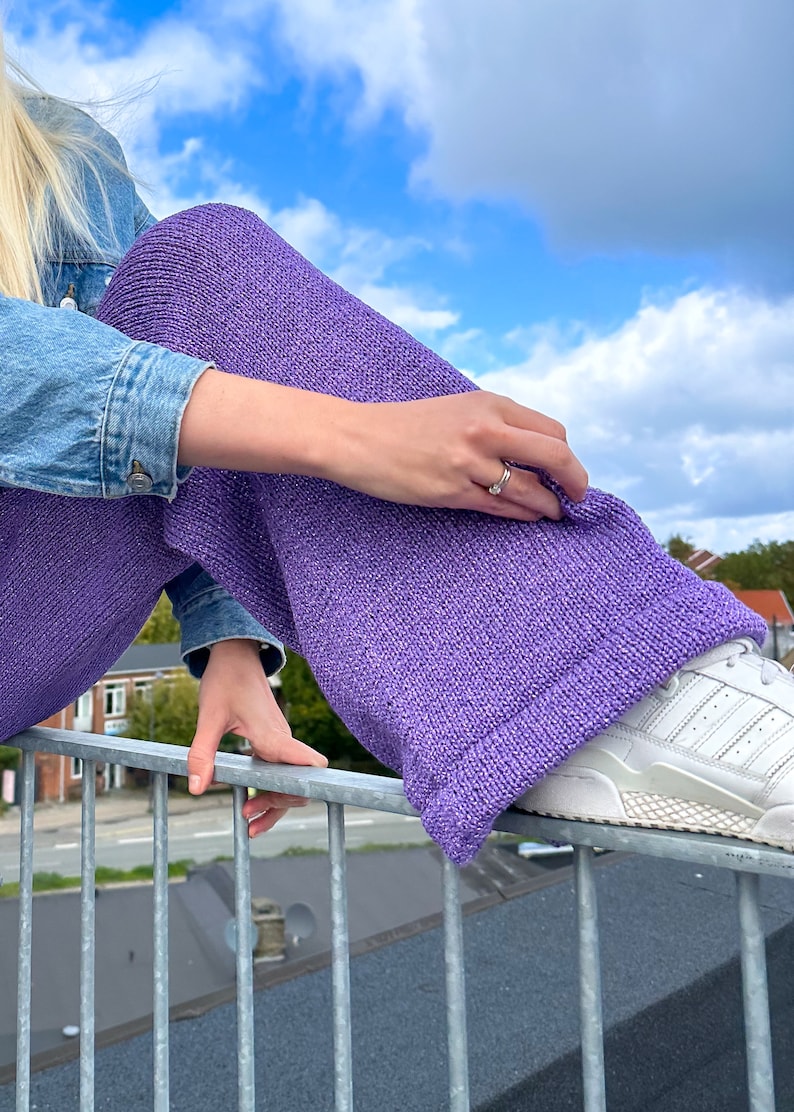 May include: A person wearing purple knitted wide-leg trousers with a denim jacket and white trainers.