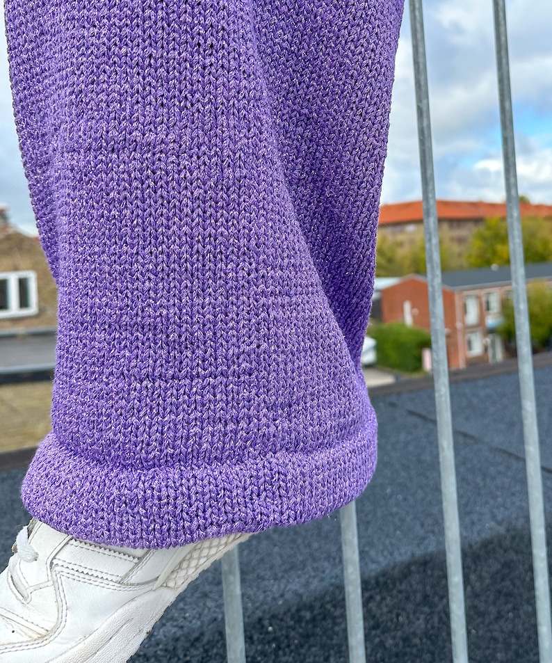 May include: Purple knitted trousers with a sparkly texture. The trousers are folded over at the bottom and are hanging on a metal railing.