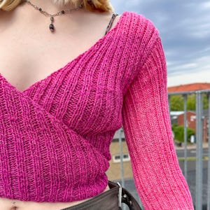 May include: A pink knit sweater with a wrap style neckline and long sleeves. The sweater is made of a soft, textured yarn.