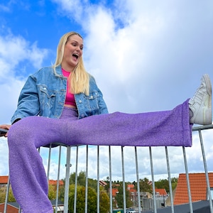 May include: A woman wearing a denim jacket, a pink crop top, and purple wide-leg trousers sits on a metal railing with one leg extended. She is smiling and looking up at the sky.
