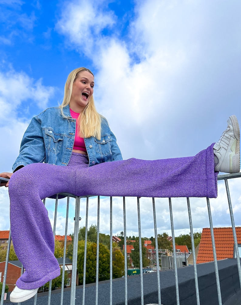 May include: A woman wearing a denim jacket, a pink crop top, and purple wide-leg trousers sits on a metal railing with one leg extended. She is smiling and looking up at the sky.
