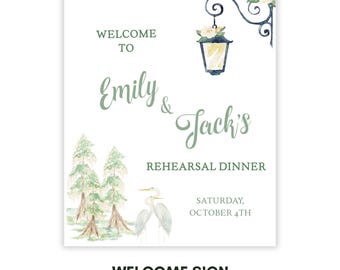 May include: A white welcome sign with watercolor illustrations. The sign reads "Welcome to Emily & Jack's Rehearsal Dinner" with the date "Saturday, October 4th." Features a lantern, trees, and birds.