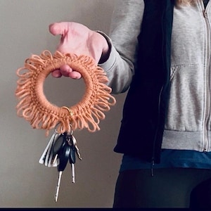 May include: A peach-colored, circular key ring with a fringe design. The key ring is holding a set of keys. The key ring is made of yarn or a similar material. The key ring is being held up in front of a light gray wall.
