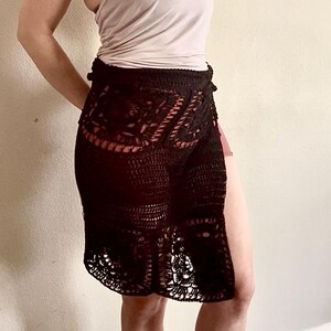 Op de afbeelding: Een zwarte gehaakte rok met een hoge taille en een zijsplit. De rok is gemaakt van een delicate, opengewerkte stof.