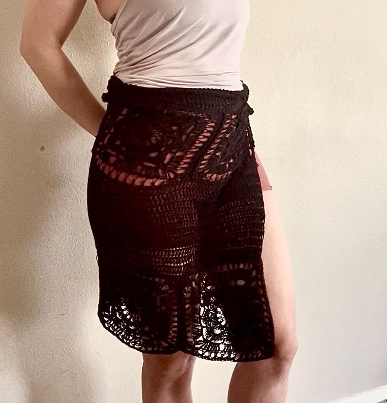 Op de afbeelding: Een zwarte gehaakte rok met een hoge taille en een zijsplit. De rok is gemaakt van een delicate, opengewerkte stof.