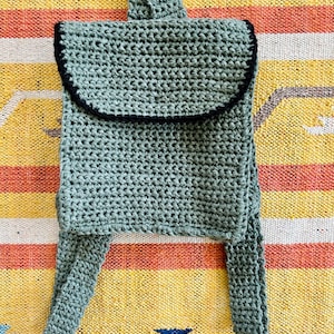 May include: A small green crocheted backpack with black trim. The backpack has a single strap and two small pockets on the front.