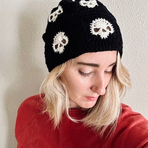 May include: A black crochet beanie with white skull designs. The beanie is being worn by a person with blonde hair.