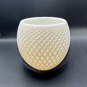 May include: A cream-colored, spherical ceramic planter with a diamond pattern. The planter sits on a black, round base. The interior of the planter is visible, and the background is black.