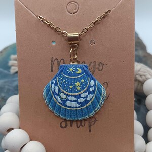 May include: A blue seashell pendant necklace with a gold chain. The pendant features a celestial design with a sun, moon, and stars. The necklace is displayed on a brown card with the words "mango shop" printed on it.