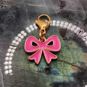 May include: A gold-coloured charm featuring a pink bow design. The bow is outlined in gold and has a glossy finish. The charm is attached to a gold-coloured clasp.