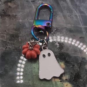 Op de afbeelding: Een Halloween-thema sleutelhanger met een regenboogkleurige sluiting. De sleutelhanger heeft een kleine, oranje pompoen bedel en een witte spook bedel met zwarte ogen. De bedels zijn met kleine metalen ringen aan de sluiting bevestigd.